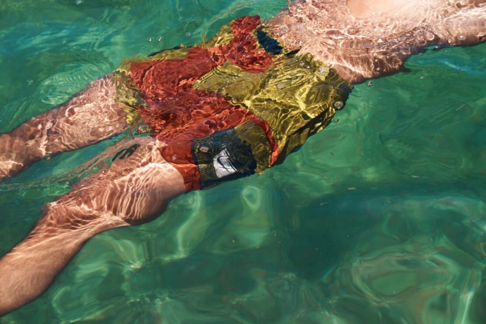 Man in patterned swim trunks partially submerged in turquoise water, viewed from above in sunlit ocean environment.
