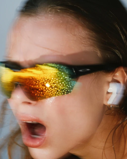 Close-up of a young adult woman shouting, wearing reflective sports sunglasses and earbuds in a brightly lit indoor setting.