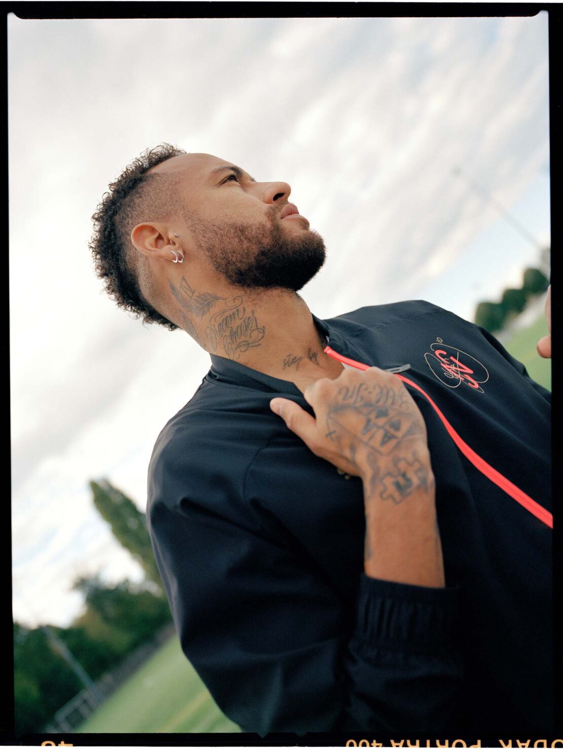 Low-angle view of a tattooed adult male with a mohawk, wearing a dark jacket with a red stripe and looking up at the sky outdoors.