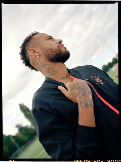 Low-angle view of a tattooed adult male with a mohawk, wearing a dark jacket with a red stripe and looking up at the sky outdoors.