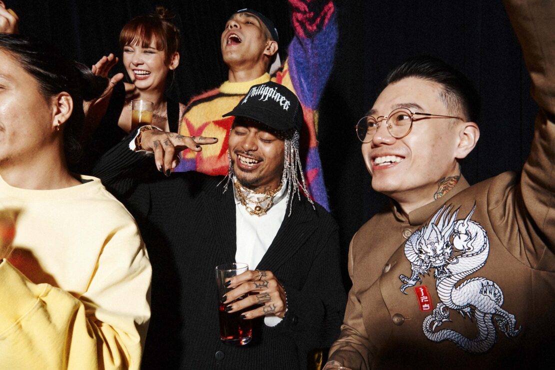 Smiling group of diverse young adults at a party, indoors with dark background, celebrating with raised arms and drinks in hand.