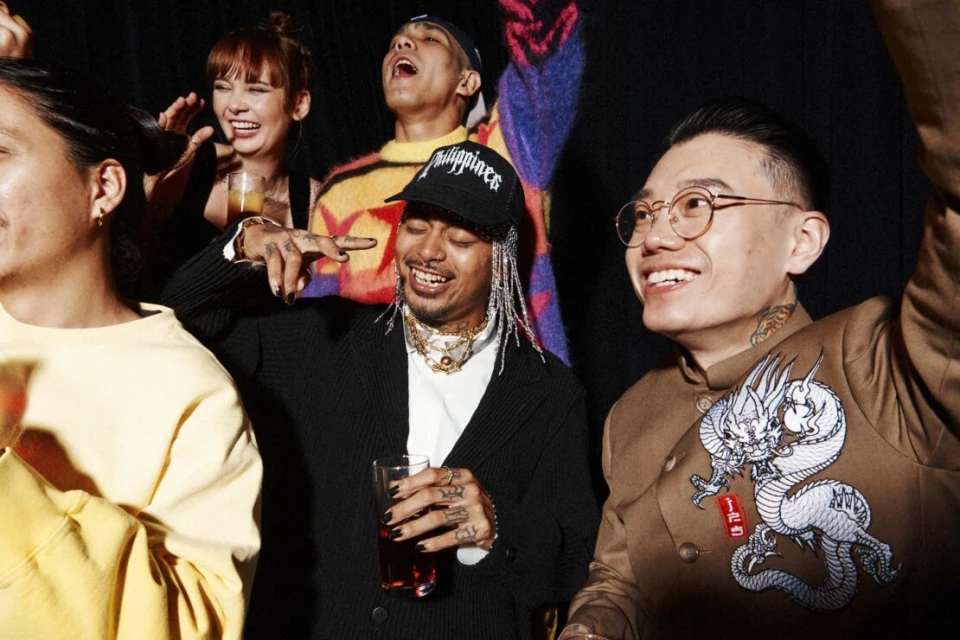 Smiling group of diverse young adults at a party, indoors with dark background, celebrating with raised arms and drinks in hand.