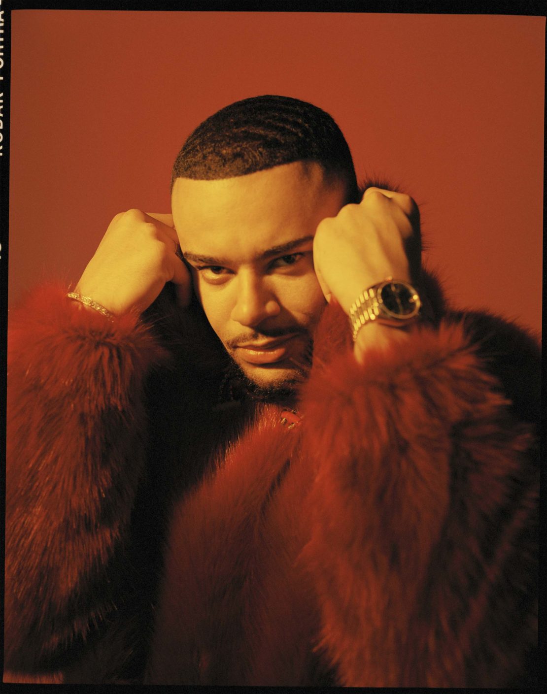 Portrait of adult male with hands near his face wearing a fur coat against an orange studio background; shot with medium framing.