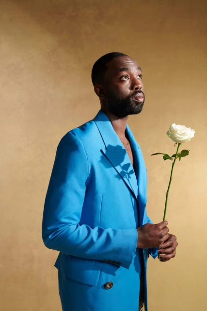 An adult Black male in a light blue suit holds a white rose, looking up and to the right, against a golden textured background.