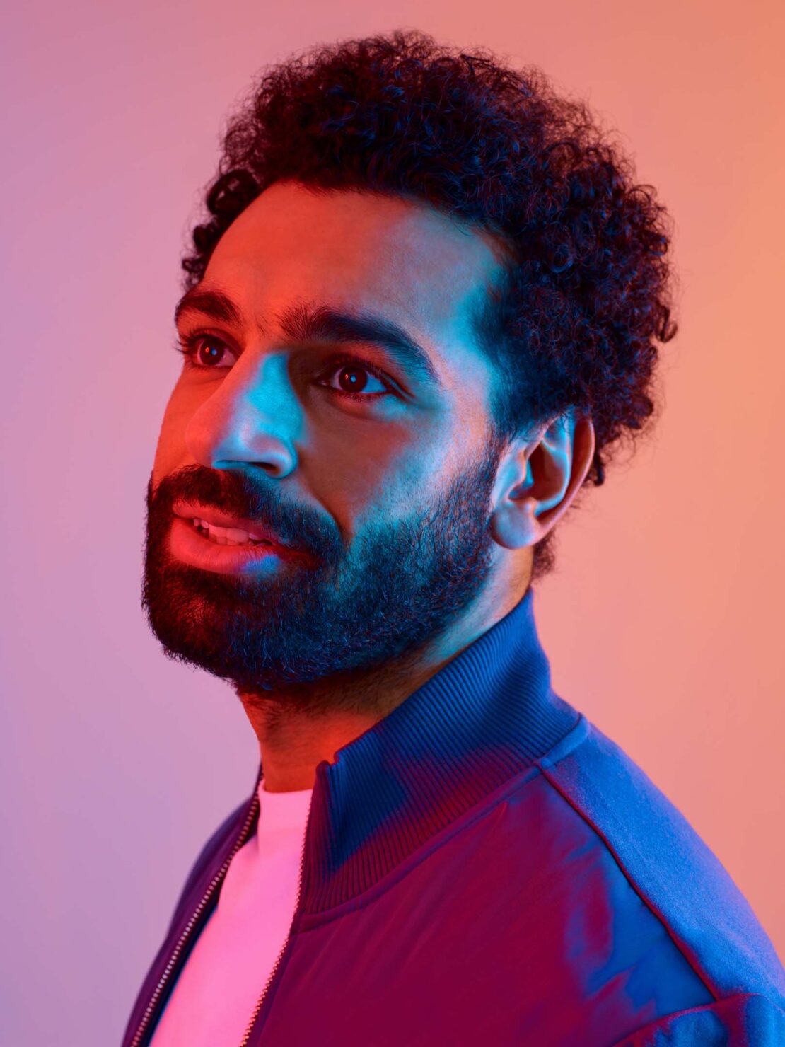 Headshot of a bearded adult male with curly hair wearing a blue jacket and a white t-shirt, gazing upwards against a multi-colored background.