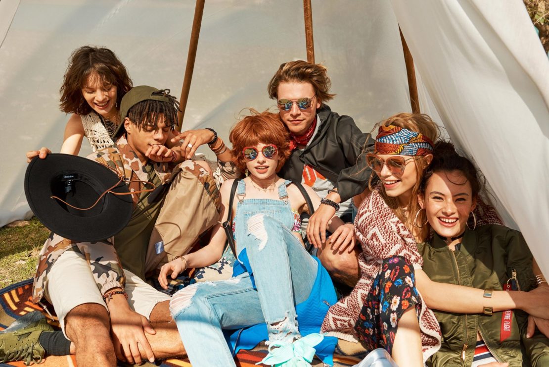 Group of six young adults gather inside a tent, wearing stylish clothing and sunglasses, sitting on a patterned blanket outdoors.