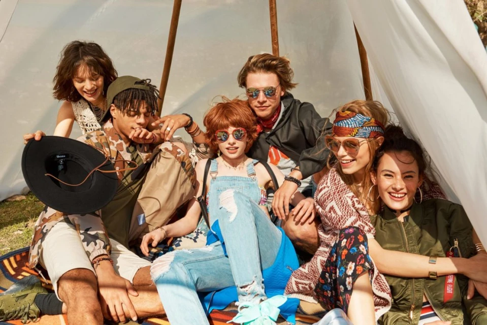 Group of six young adults gather inside a tent, wearing stylish clothing and sunglasses, sitting on a patterned blanket outdoors.