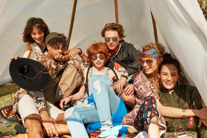Group of six young adults gather inside a tent, wearing stylish clothing and sunglasses, sitting on a patterned blanket outdoors.