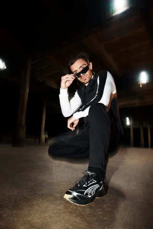 Man in sunglasses and athletic apparel poses squatting in a dimly lit industrial space, showcasing fashion sneakers.