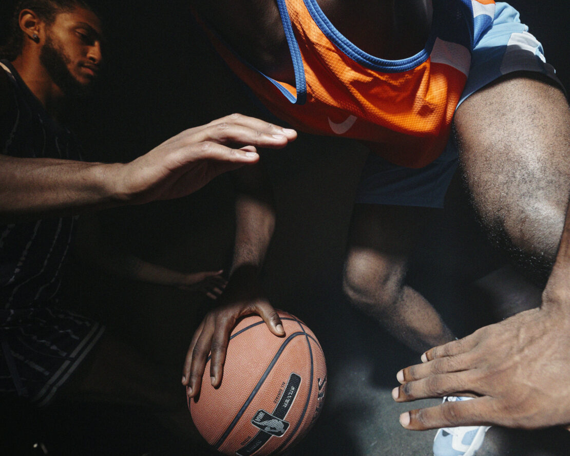 Two basketball players vie for the ball in a dramatically lit indoor game; orange and blue team jerseys are visible against a dark backdrop.