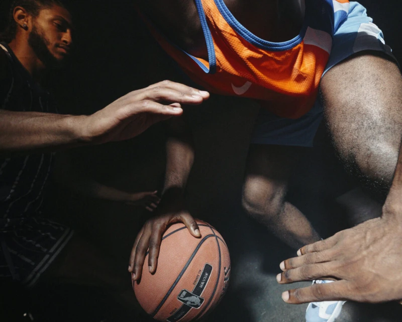 Two basketball players vie for the ball in a dramatically lit indoor game; orange and blue team jerseys are visible against a dark backdrop.