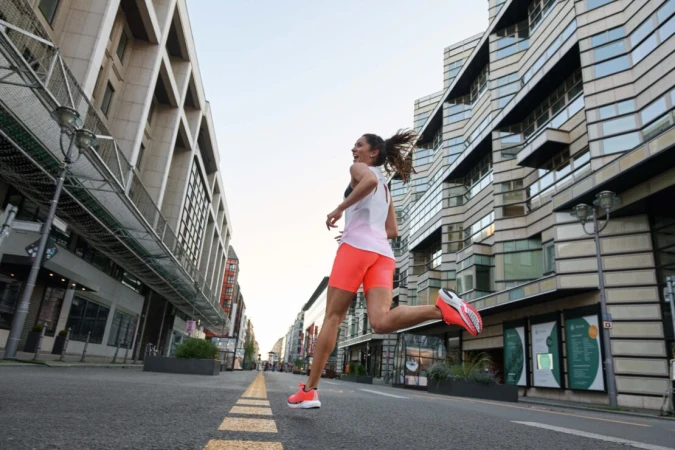 A woman in sportswear runs on an empty city street with tall buildings, captured from a low angle, highlighting fitness and urban environment.