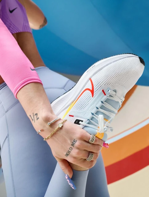 Close-up of an adult wearing sportswear holding a white Nike running shoe with a red swoosh, featuring tattoos, bracelets, and rings.