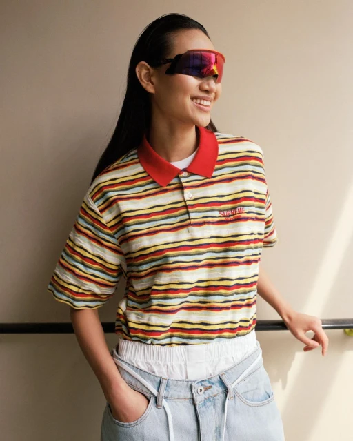 Smiling Asian woman wears striped polo shirt, retro sunglasses, jeans against neutral wall. Casual fashion with 90s style aesthetic.