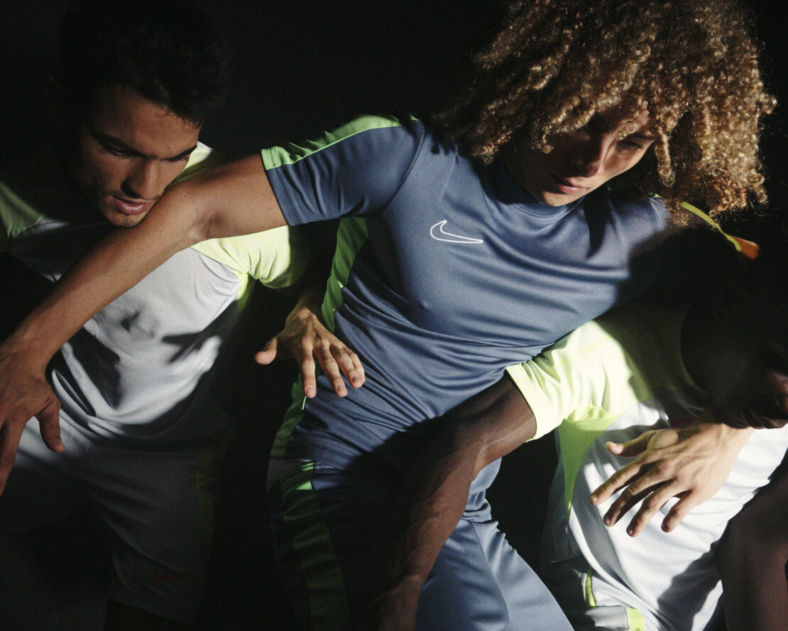 Three athletes wearing athletic clothing stand close together in a studio with dark background, illuminated by studio lighting.