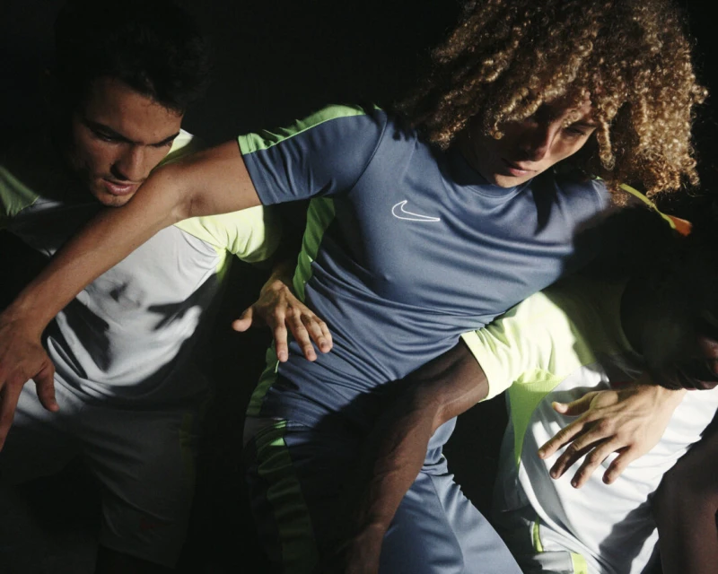 Three athletes wearing athletic clothing stand close together in a studio with dark background, illuminated by studio lighting.