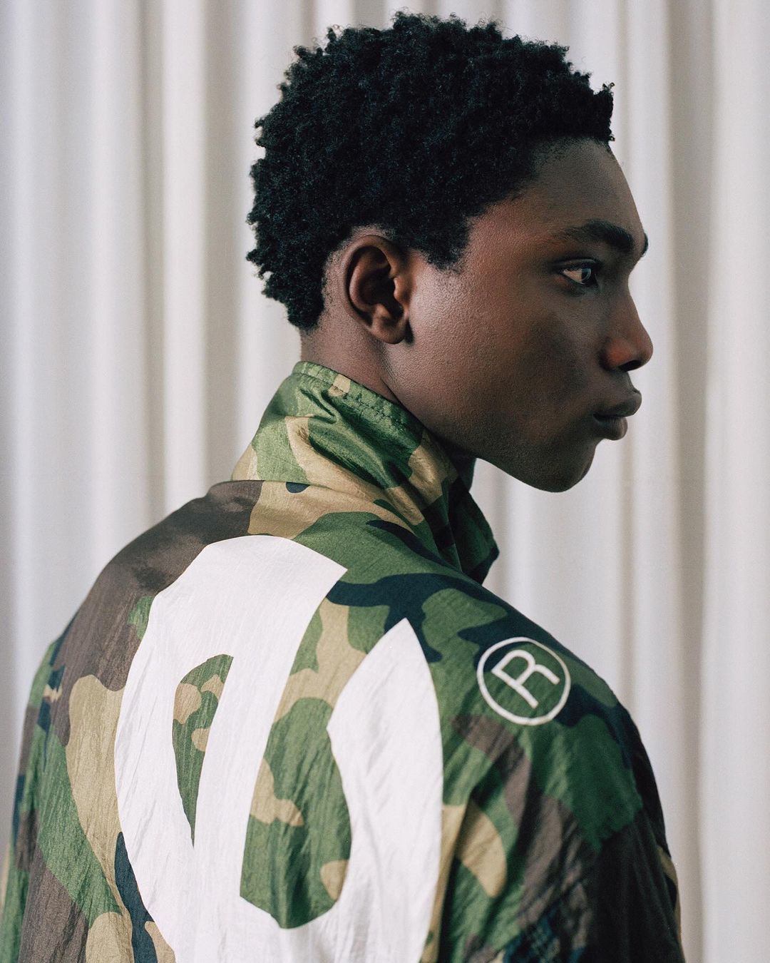 Studio shot of a Black male model in a camouflage-patterned jacket, looking off-camera. The jacket features a large white graphic.