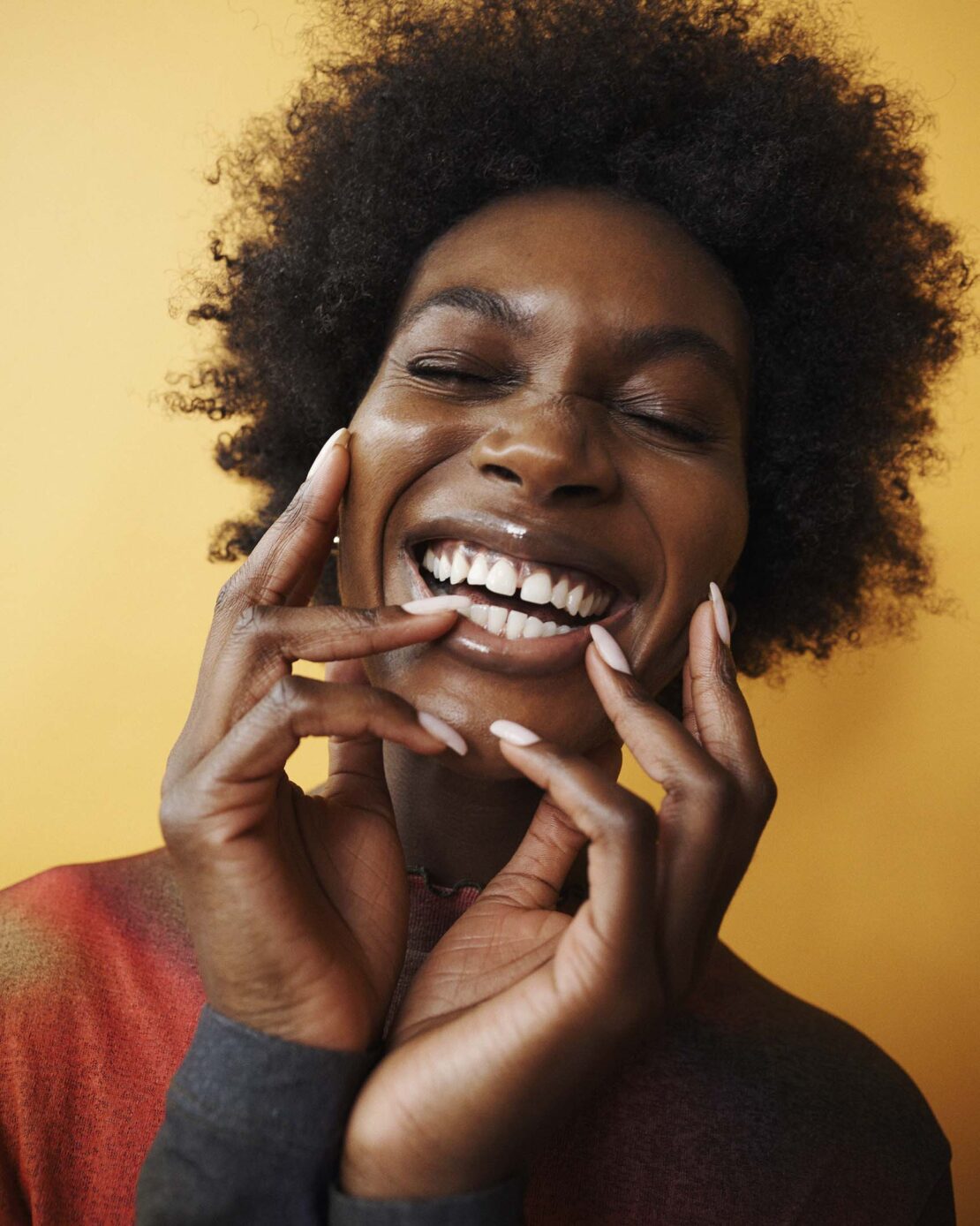 Portrait of a smiling Black woman with dark curly hair, eyes closed, pale pink nails, wearing a colourful top against yellow background.