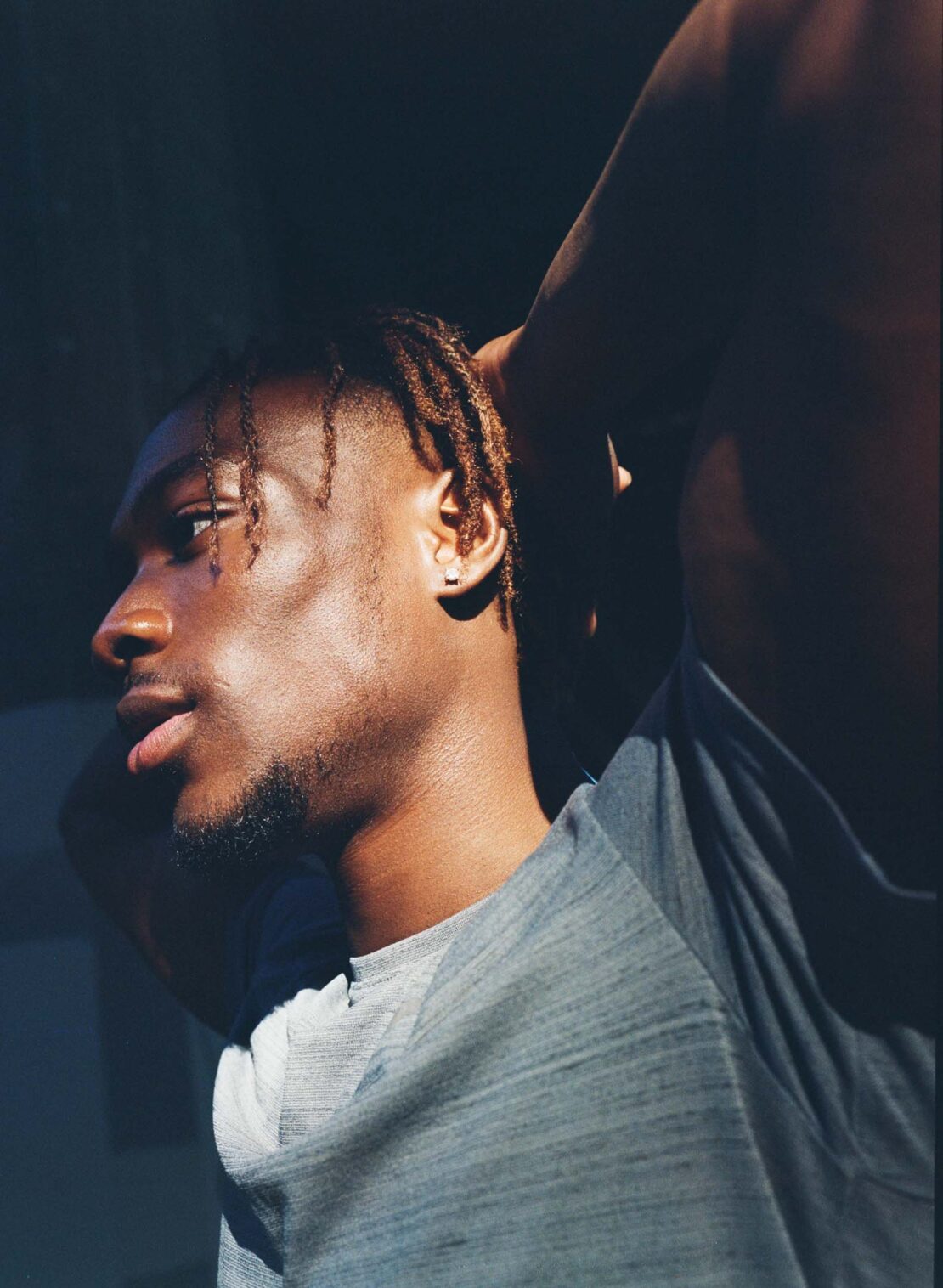 Portrait of a Black man in a gray t-shirt, hands behind his head, standing indoors against a dark background with sunlight on his face.