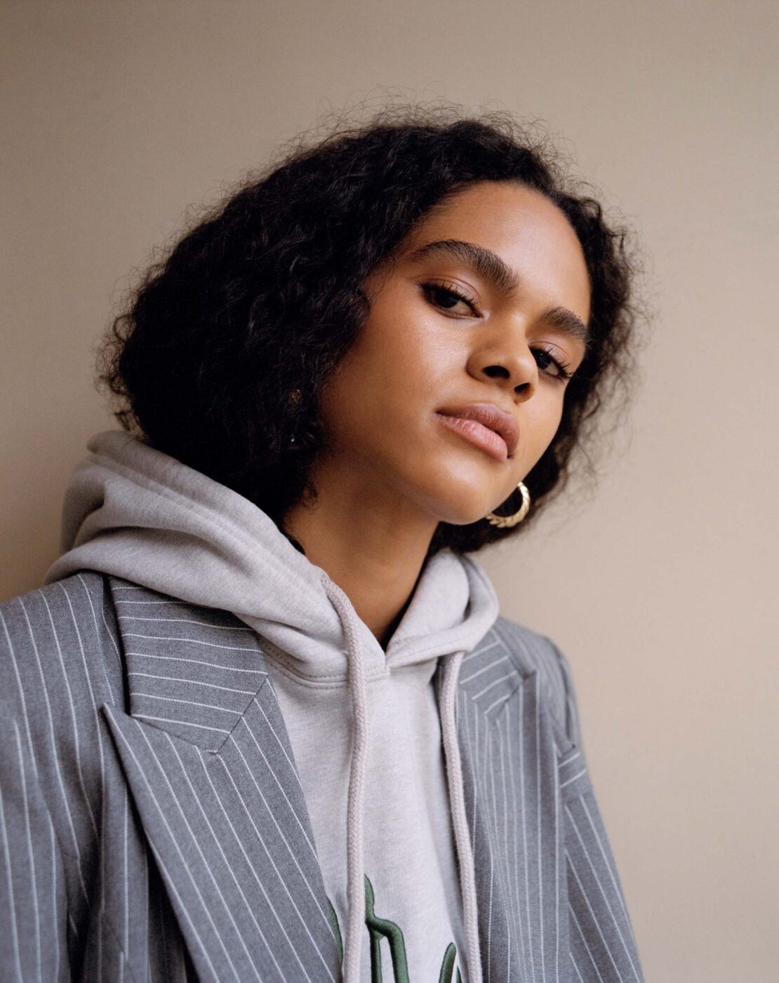 Portrait of a young woman with curly dark hair wearing a pinstripe blazer over a hoodie in a neutral indoor setting. Fashion shot.