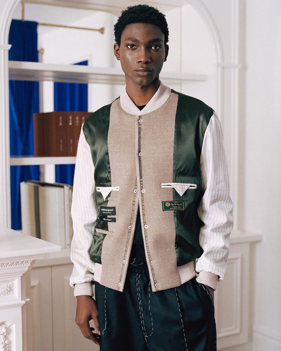 A stylish young man poses indoors wearing a color-blocked bomber jacket with black pants and white stitching, standing in front of a bookcase.