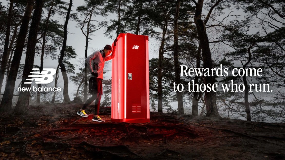 An athletic man pauses by a red New Balance branded locker on a forest trail with the tagline 'Rewards come to those who run'.