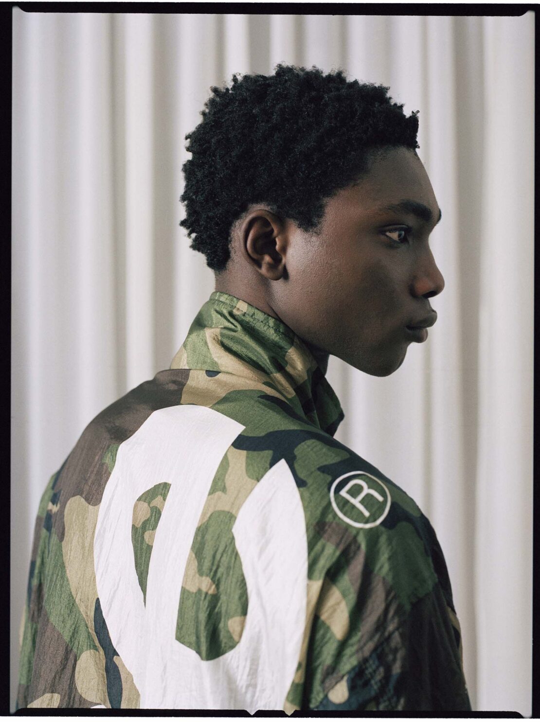 Portrait of an adult man in profile wearing a camouflage jacket with a white logo, set against a neutral-toned curtain backdrop indoors.