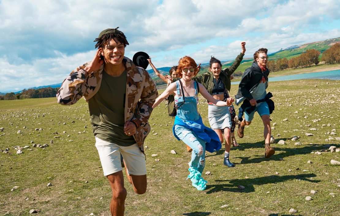 Group of young friends running across a grassy field near a body of water under a bright sky