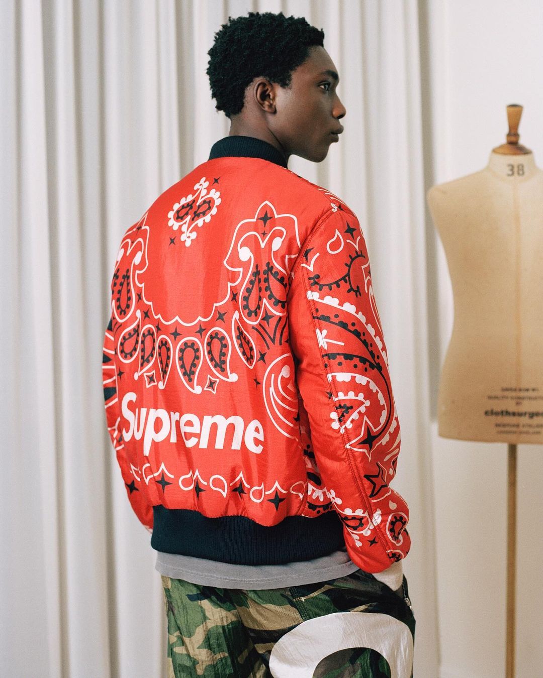 A young man is seen from the back and side, wearing a red paisley-print Supreme jacket, standing indoors near a mannequin.