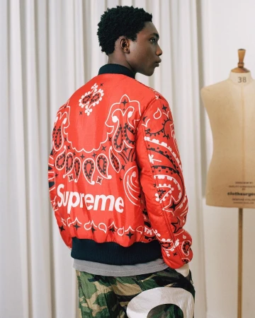 A young man is seen from the back and side, wearing a red paisley-print Supreme jacket, standing indoors near a mannequin.