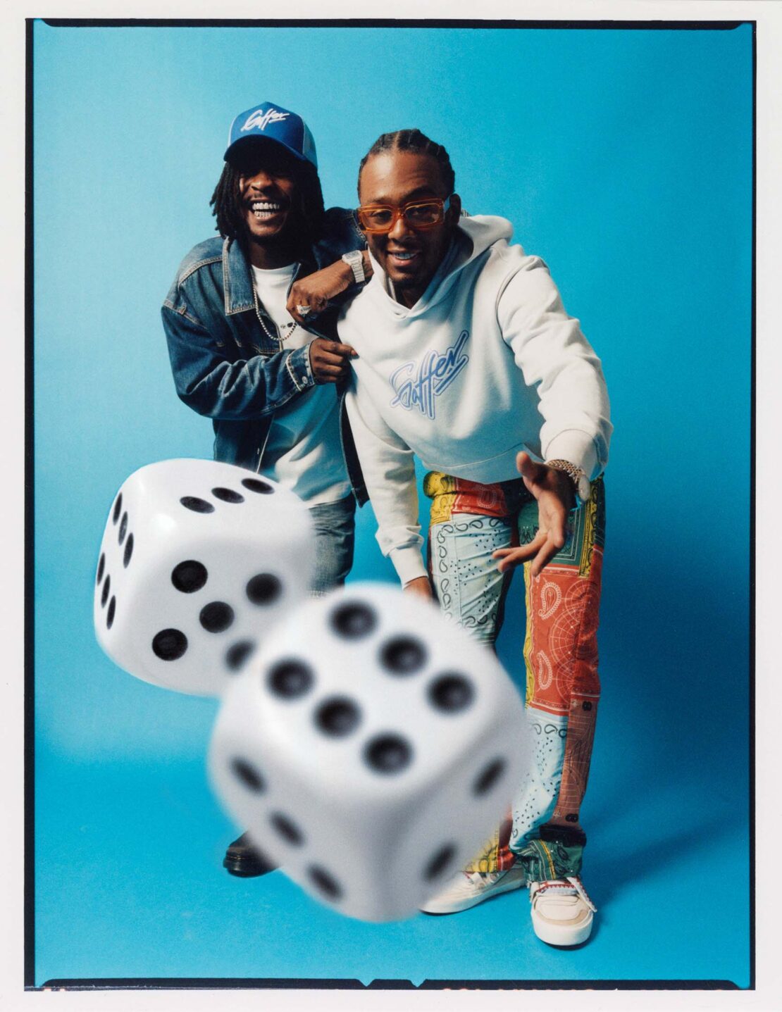 Two young adult men pose indoors wearing stylish casual clothes with large dice in front of a vibrant turquoise background.