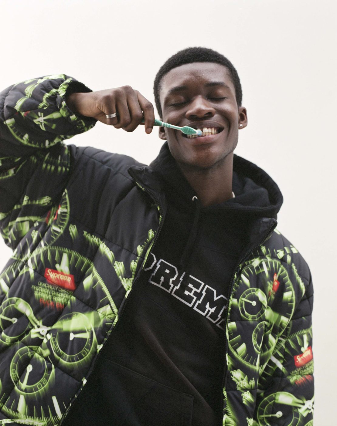 A smiling Black man in a patterned jacket and hoodie brushes his teeth with a green toothbrush in an indoor environment.