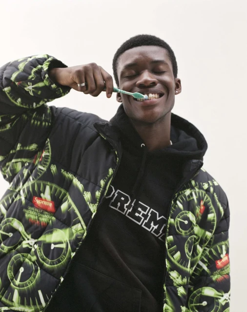 A smiling Black man in a patterned jacket and hoodie brushes his teeth with a green toothbrush in an indoor environment.
