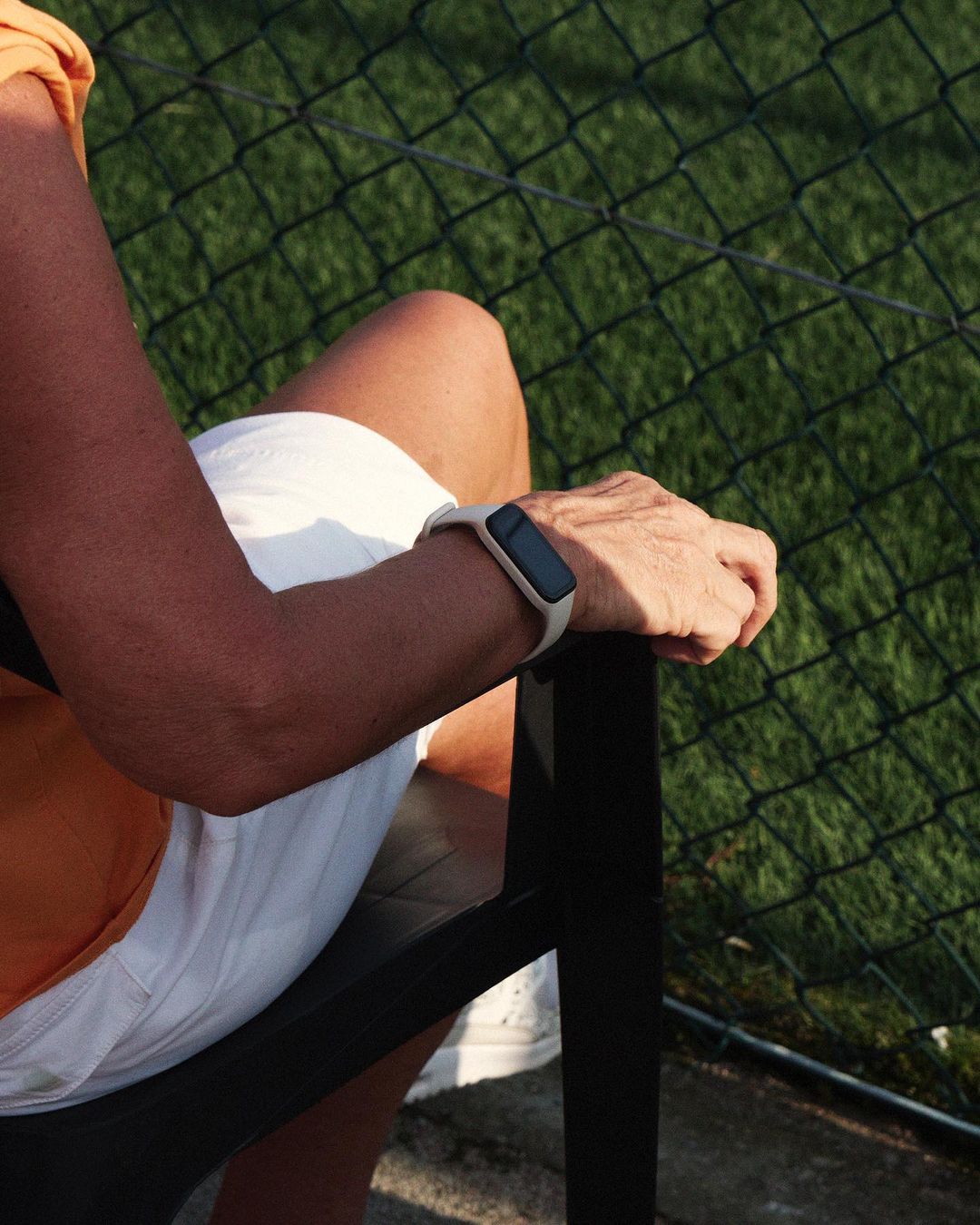 An adult sits in a black chair, wearing a fitness tracker, white shorts, and an orange shirt, with a black chain-link fence in the background.