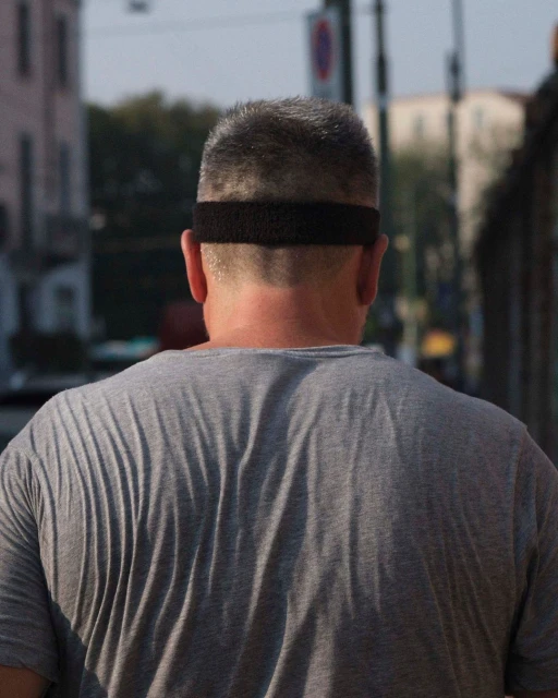 Back view of an adult male in a gray t-shirt and black headband, walking on a city street with buildings and trees in the background.