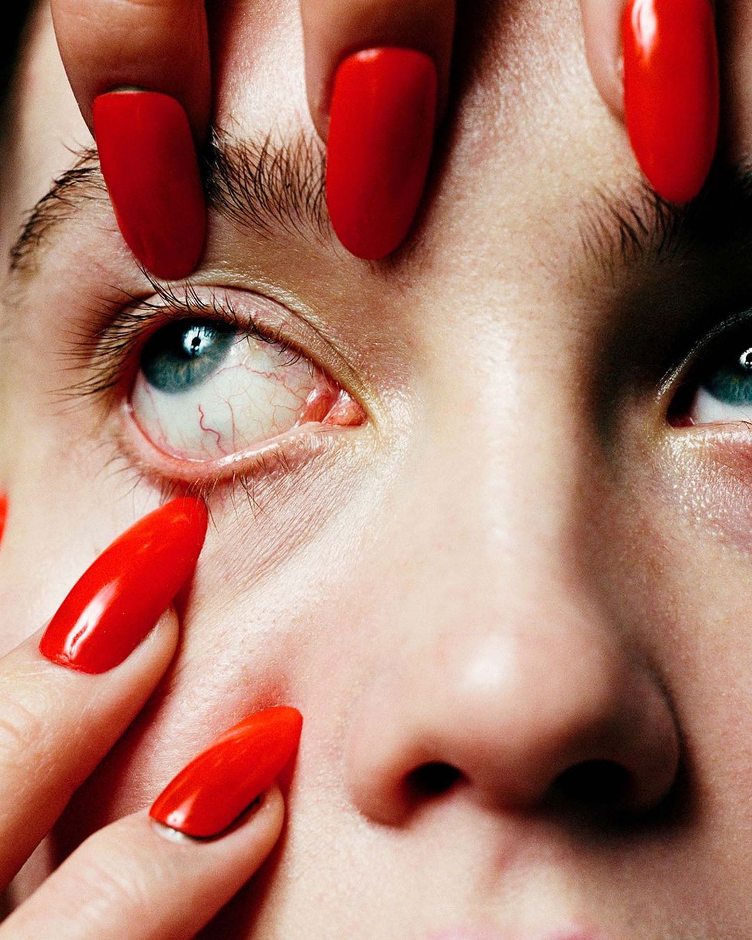 Close-up of a face with light eyes surrounded by fingers with long, glossy red nails, creating a visually striking beauty image.