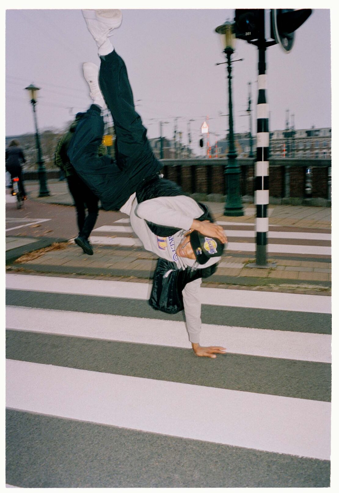 Person does a handstand on a crosswalk in an urban setting with street lights, buildings, and a pedestrian walking by in the background.