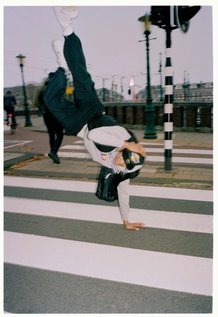 Person does a handstand on a crosswalk in an urban setting with street lights, buildings, and a pedestrian walking by in the background.