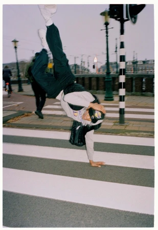 Person does a handstand on a crosswalk in an urban setting with street lights, buildings, and a pedestrian walking by in the background.