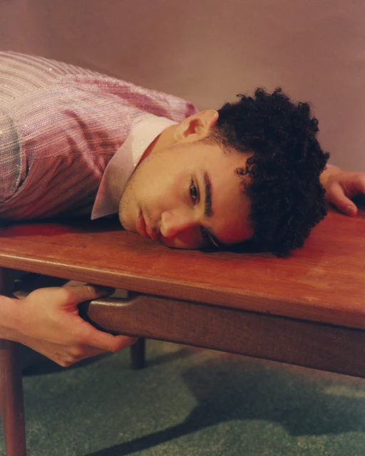 Man in shimmering pink shirt rests head on wood table indoors, lit with warm color grading. Calm expression, eye-level angle.