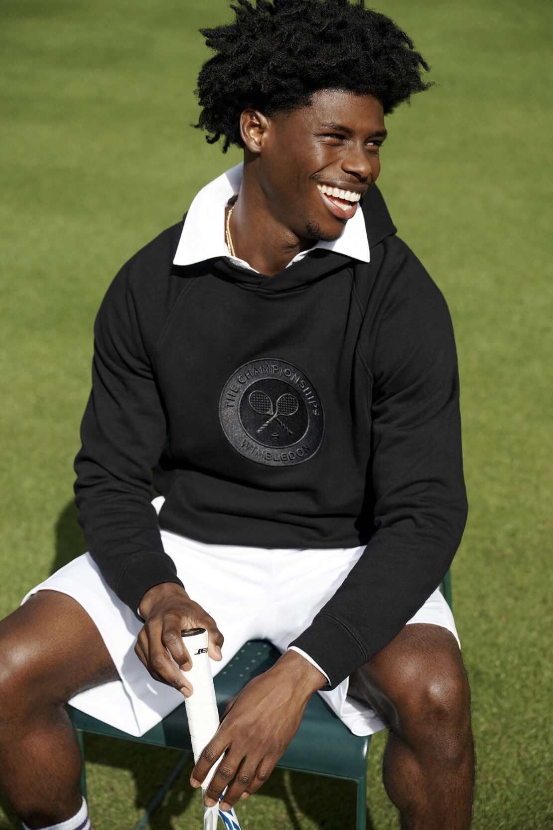 A smiling Black man wearing a black sweatshirt with the Wimbledon logo sits on a chair in front of a green lawn, holding a tennis racket.