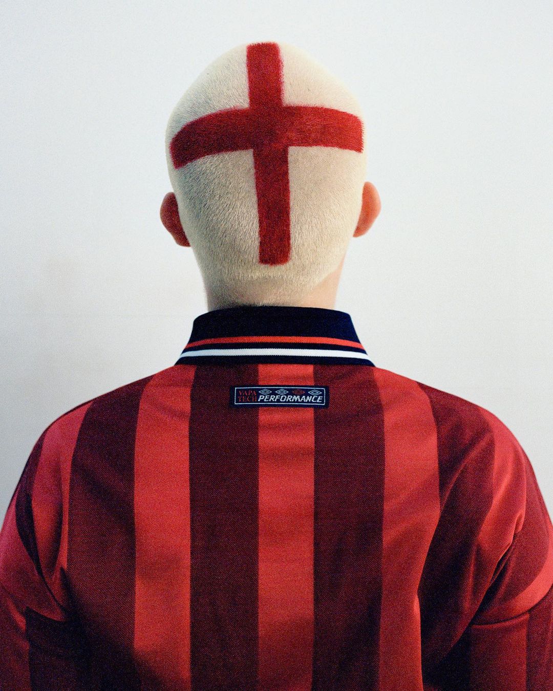 Back view of person with a cross shaved into their blonde hair, wearing a red and black striped collared shirt against a white wall.