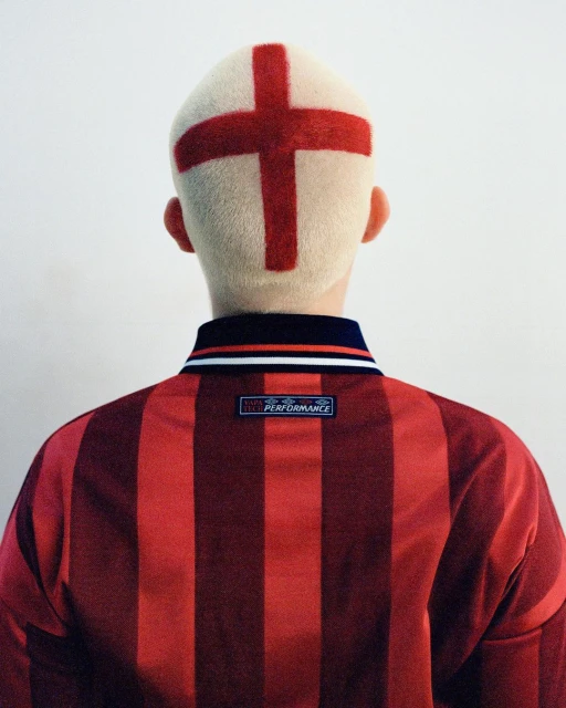 Back view of person with a cross shaved into their blonde hair, wearing a red and black striped collared shirt against a white wall.