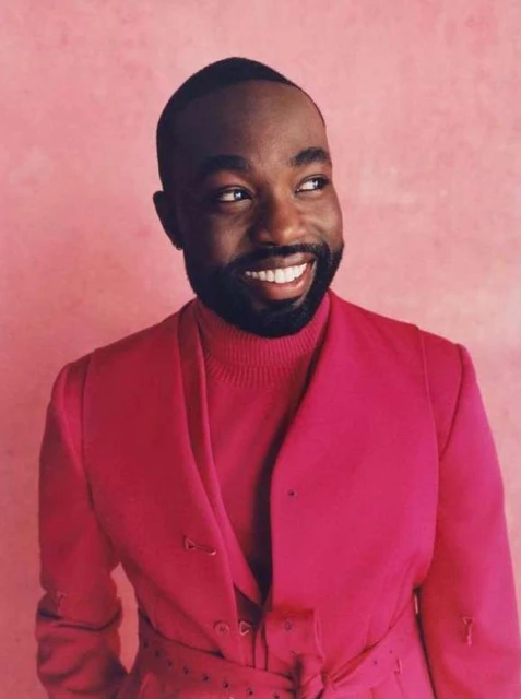 Portrait of handsome smiling Black man in pink turtleneck and pink suit jacket looking off to the side, set against a pink background.