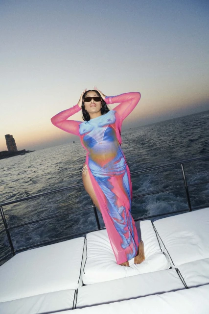 Adult woman in printed dress and sunglasses poses on a boat at sea with a distant stone structure visible on the horizon at dusk.