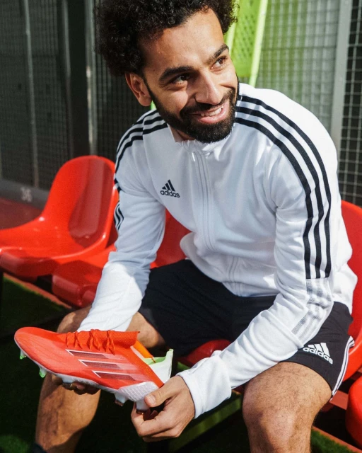 An adult male smiles while holding an orange Adidas soccer shoe; he wears Adidas shorts and jacket near a stadium bench.