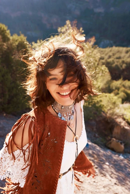 Smiling woman with bohemian style wearing a suede fringe vest and necklace in a natural outdoor setting with sunlight and trees.