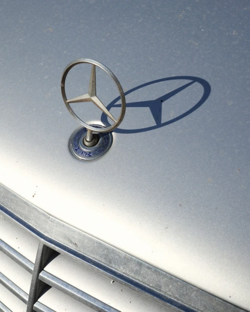 Close-up of a Mercedes-Benz hood ornament on a silver car, showing the circular star logo and the car's front grill.