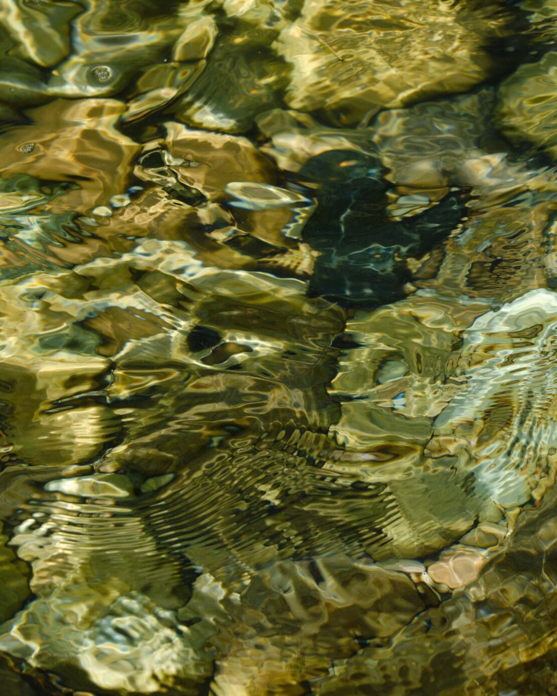 Rippled water covers multicolored rocks, creating a mesmerizing natural abstract pattern with brown, green, and black tones.