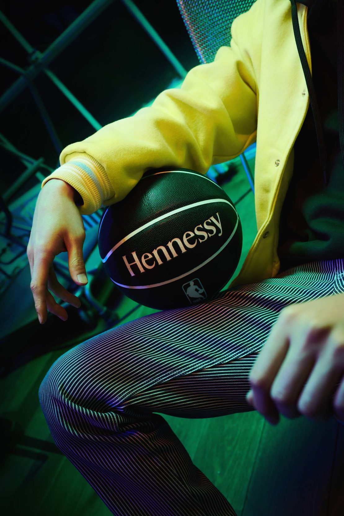Person sits with a branded Hennessy basketball, wearing a yellow jacket and striped pants, under cool green and blue lighting.